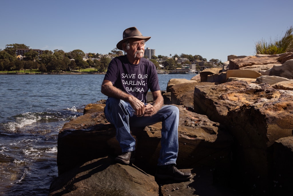 A person wearing jeans, t-shirt, boots and hat sits on rocks by the water.