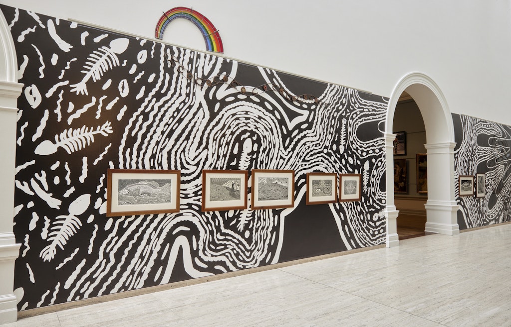 A large black-and-white print forms a background on a gallery wall on which framed artworks are hung.