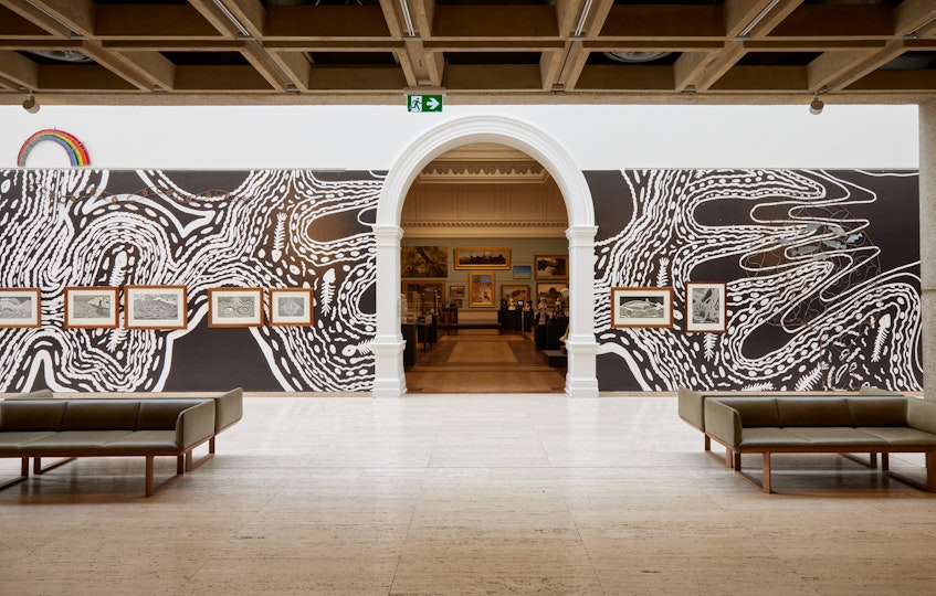 A large black-and-white print forms a background on a gallery wall on which framed artworks are hung.