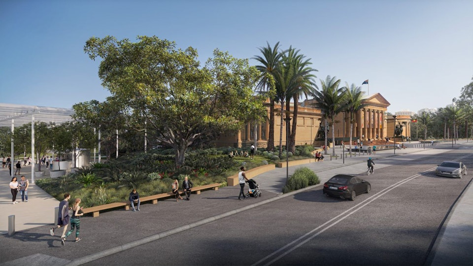 A sandstone building with a colonnaded portico with pedestrian paths and a roadway. To its left are landscaped gardens and a plaza.