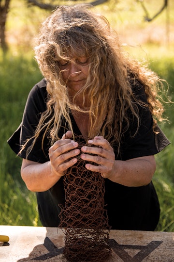 A person with long hair dressed in black stands outdoors at work surface with both hands wrapped around a column of wire mesh.