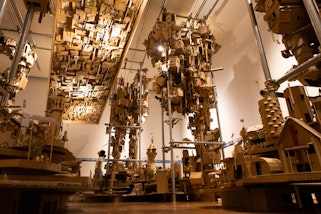 A person stands within a gallery space surrounded by complex structures made up of small cardboard buildings.