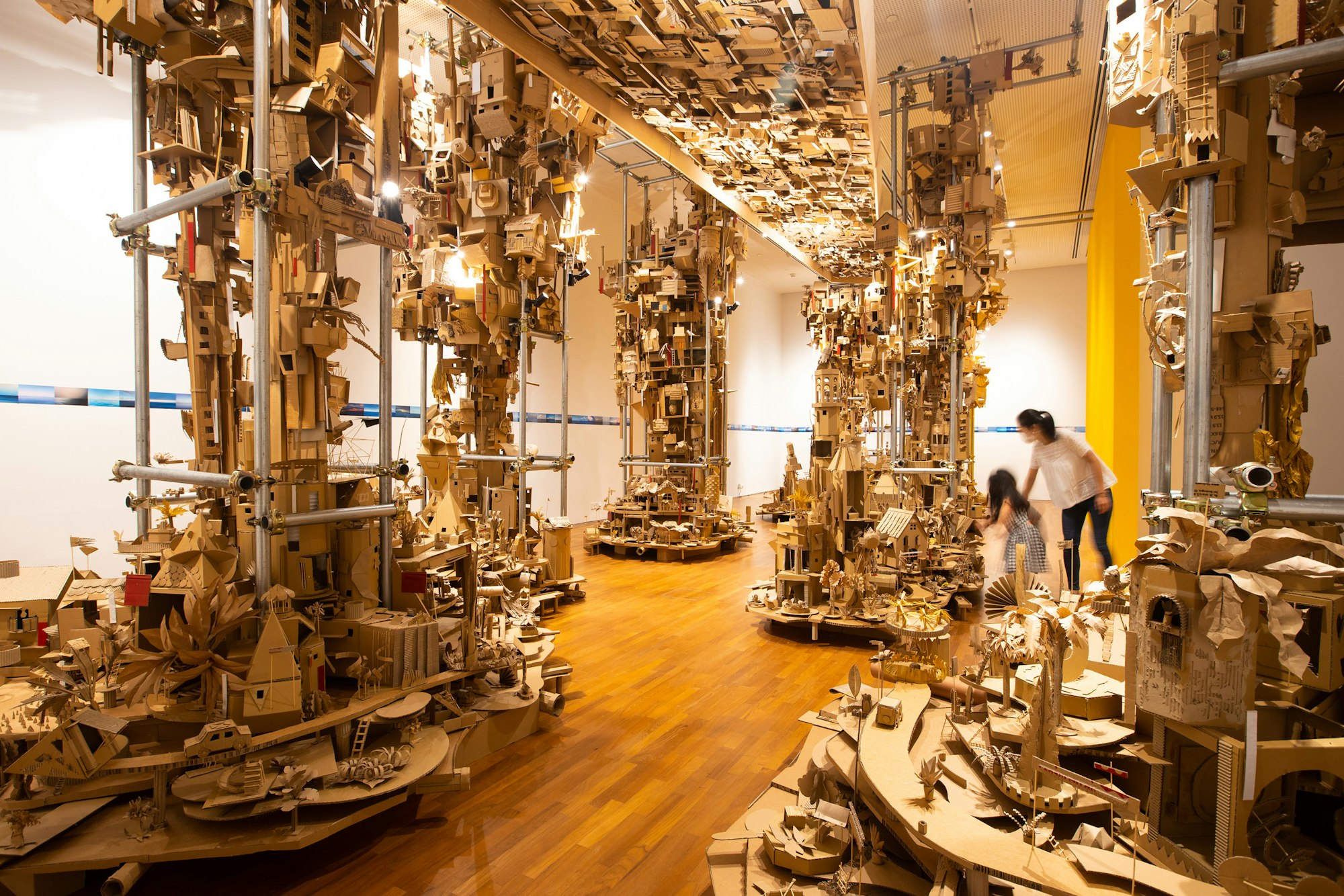 An adult and child stand within a gallery space surrounded by complex structures made up of small cardboard buildings.