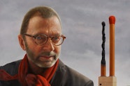 A short-haired person with glasses and a red scarf in front of a sculpture of giant matchsticks