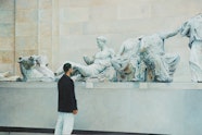 A short-haired person wearing white pants and a black jackets stands in front of a museum display of classical sculptures
