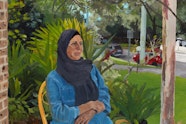 A person wearing a headscarf, long top and pants sits on a chair on a verandah with gardens, cars and a street behind them