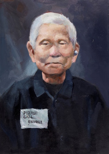 The head and torso of a person with short grey hair wearing a hearing aid and black top on which a sewn-on label reads 'Please call 96774811'