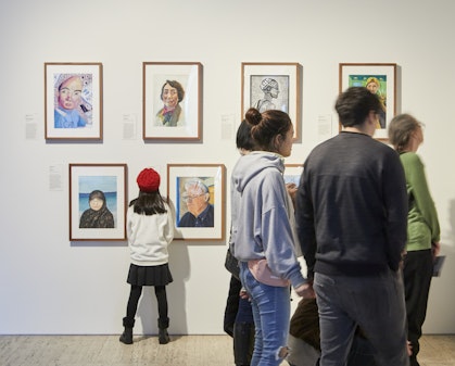 A child in a red beret reads a label on a gallery wall as other people look at the rows of framed portraits on display