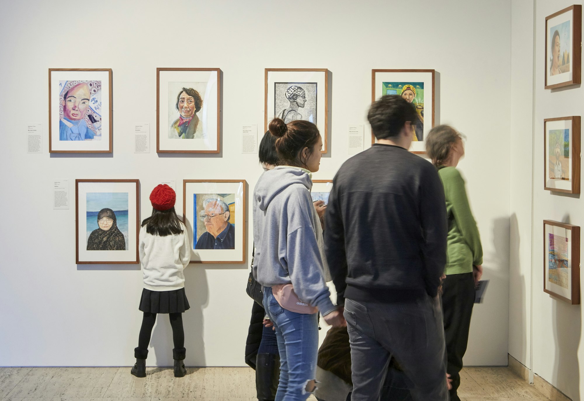 A child in a red beret reads a label on a gallery wall as other people look at the rows of framed portraits on display