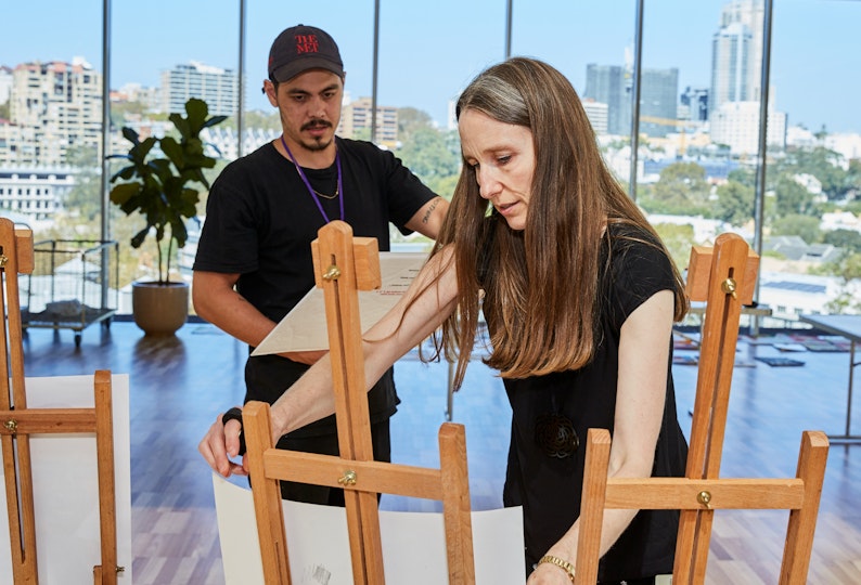 A person in a cap holds a piece of paper while another long-haired person props a piece of paper on an easel