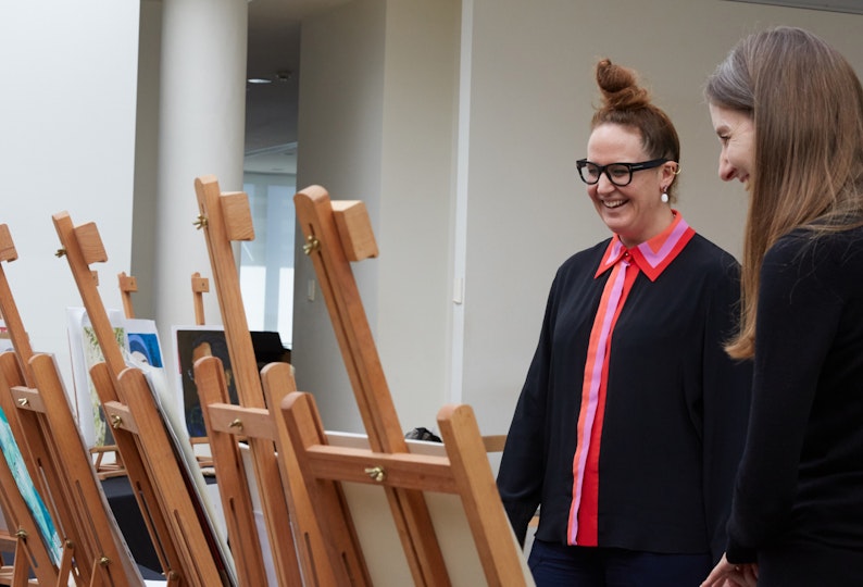 A person with glasses and hair in a top knot laughs with a long-haired person while they look at artworks on easels
