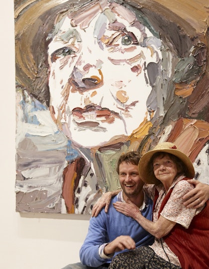 Two people sit with their arms around each other in front of a large painting of one of them