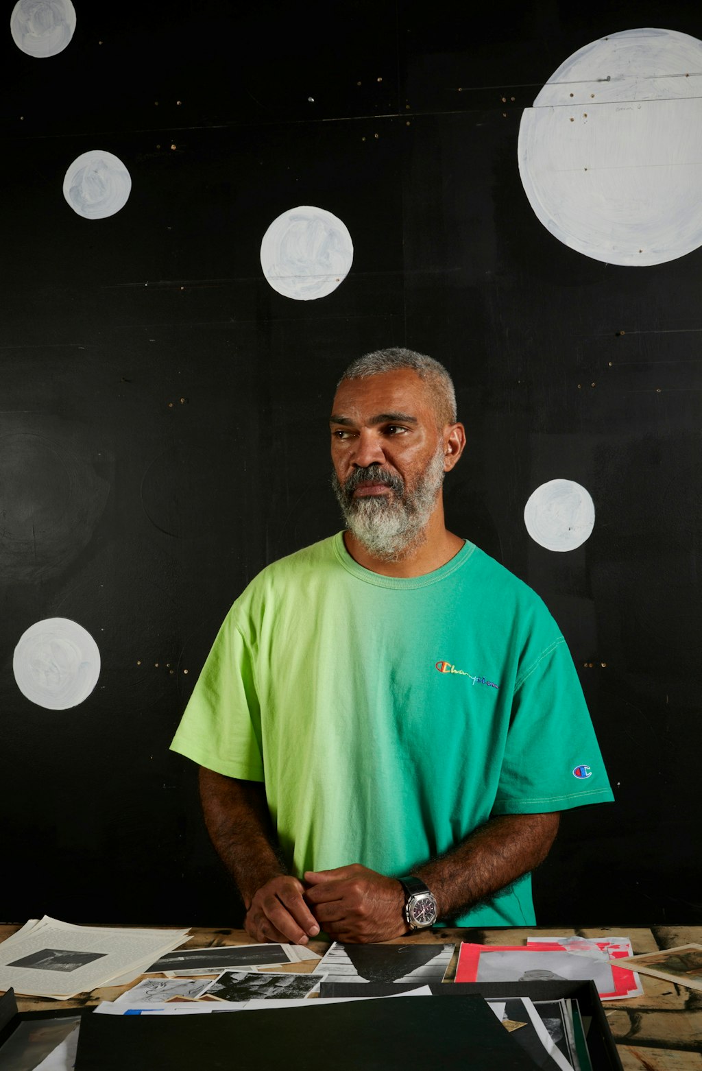 A bearded, short-haired person in a t-shirt at a table on which lies multiple pieces of paper with images. The background is black with white dots of various sizes.