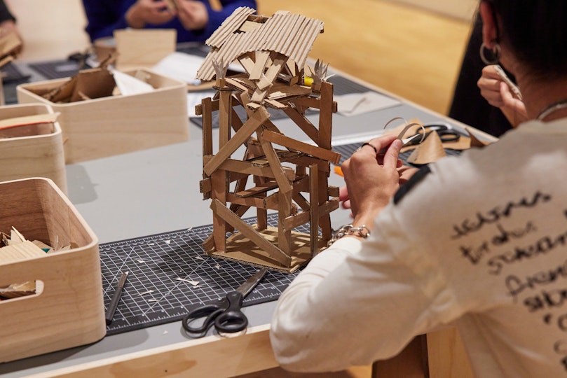 A person sits at a table working on a small cardboard structure