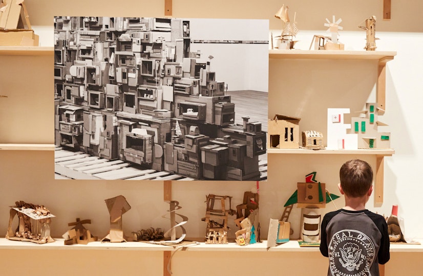 A child stands looking at small cardboard structures on shelves. A black-and-white photo of a complex cardboard structure hangs in front of the shelves.