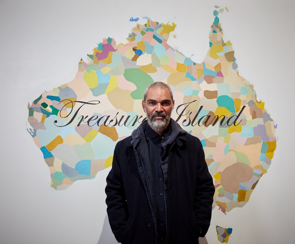 A bearded, short-haired person stands in front of a image of Australia divided into multi-coloured sections on which 'Treasure Island' is written