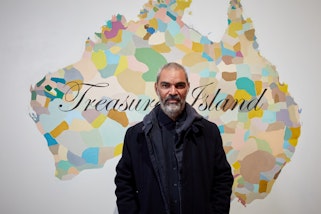 A bearded, short-haired person stands in front of a image of Australia divided into multi-coloured sections on which 'Treasure Island' is written