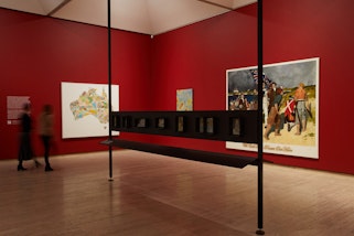 Photograph of an exhibition space with three paintings on the wall and a display shelf in the middle.