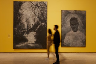 Photograph of an exhibition space with warm yellow walls and two large paintings hung in portrait format. Two people walk in the middle of the room.