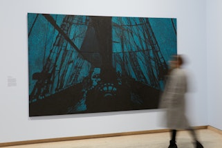 A photograph of a large painting in an exhibition space with light blue walls. A person moves on the right of the room.