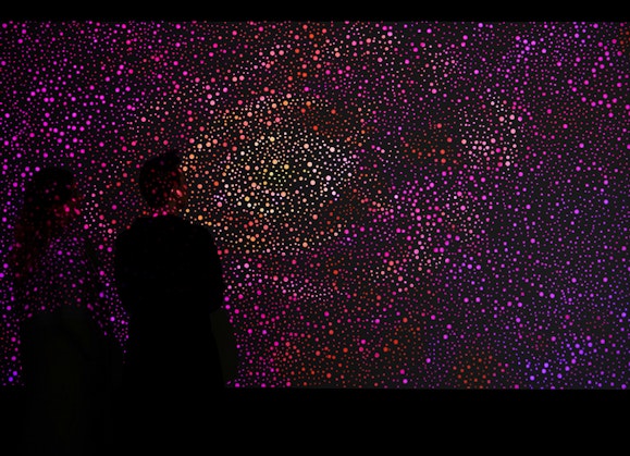 Photograph of two people standing in front of a video artwork. The artwork shows many dots of magenta, red, white and yellow on a dark background.
