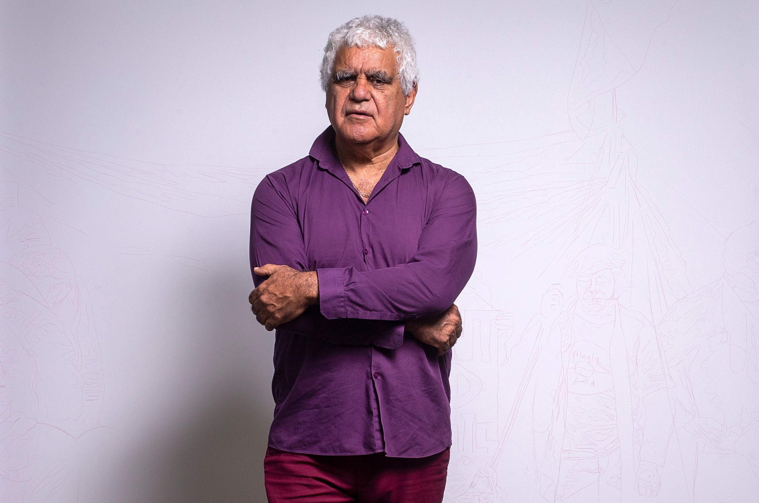 A short-haired person wearing a purple shirt and maroon pants stands with their arms crossed