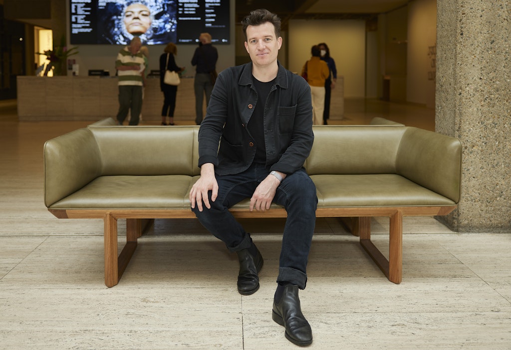 A short-haired person sits in a gallery space on a bench seat that has wooden legs and olive green leather upholstery