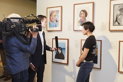 A person stands in front of a row of framed portraits while being interviewed by a person with a microphone and filmed by a person with a camera