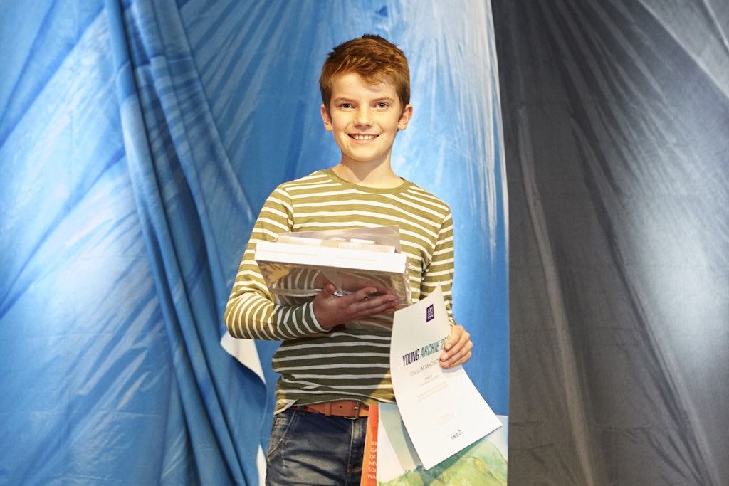 A young person in striped t-shirt stands holding a bundle and a certificate with the words 'Young Archie'