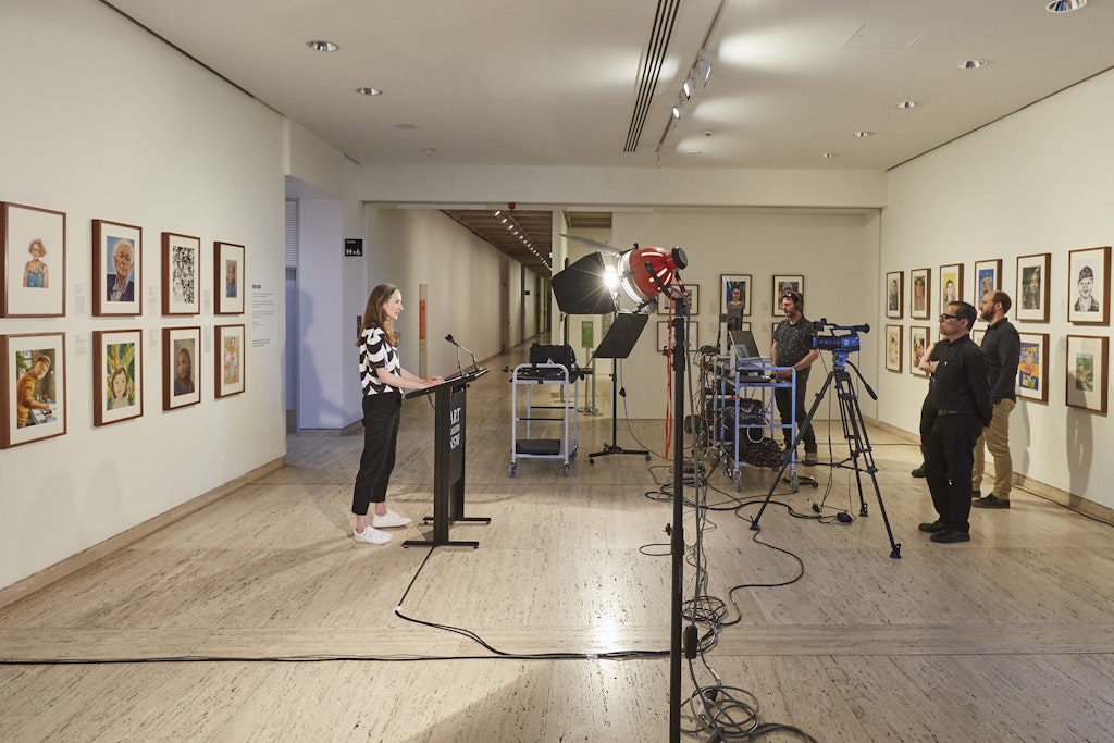 A person stands at a lecturn in front of audio-visual equipment and four other people within a gallery space hung with framed portraits