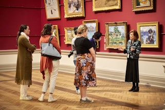Six people in a gallery space in front of a wall of gold-framed paintings