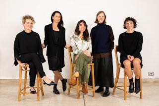 Five women in a row. Three are sitting on stools and two are standing.