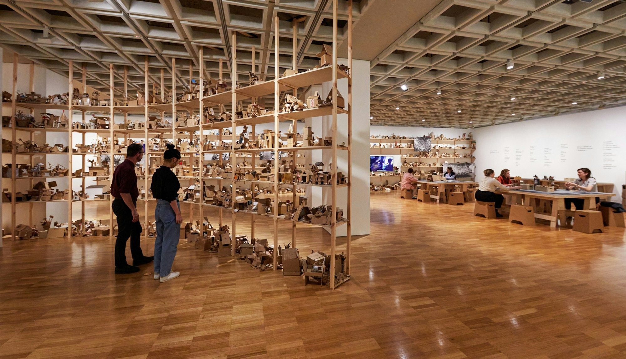 Two people view shelves full of small cardboard structures while groups of people sit at large tables making other cardboard structures