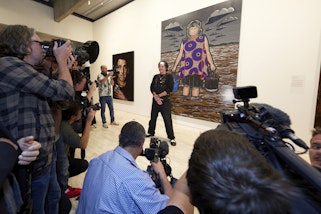 A group of people with cameras pointing at a  person standing in front of a large painting in a gallery space