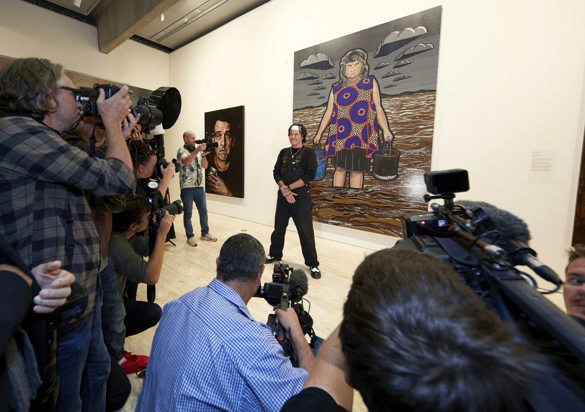 A group of people with cameras pointing at a  person standing in front of a large painting in a gallery space
