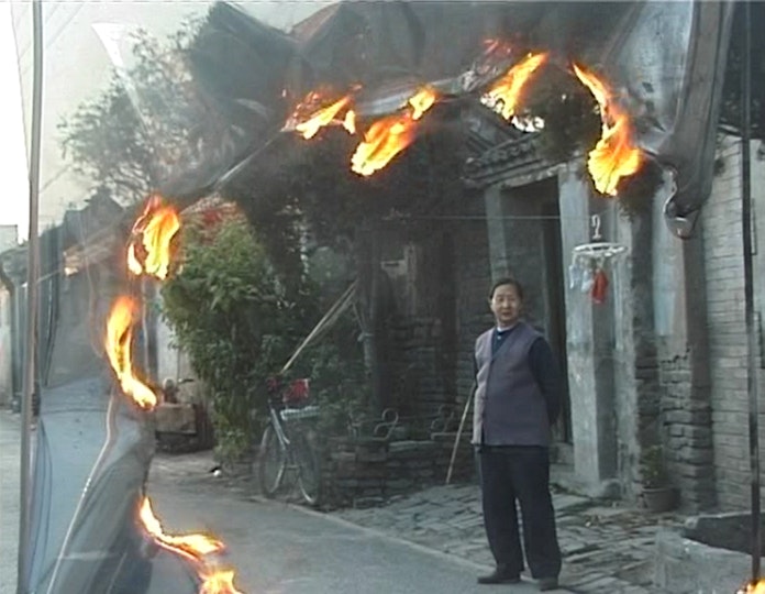 A person stands in a street in front of a row of small brick buidlings. There is a half-circle of flames on the left and top of the image.
