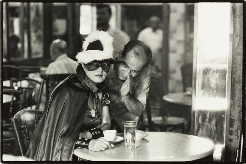A masked and caped person sits at a cafe table while another person leans over near them with their hand held to their face as if whispering