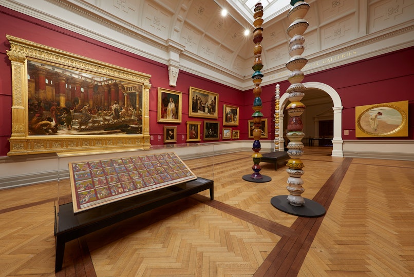 Paintings in ornate gold frames hang in a red-walled gallery which also houses pole-like sculptures and a painting within a showcase
