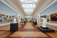 A large gallery space with a moulded ceiling with vaulted skylight and blue walls hung with many paintings. Sculptures and display cases stand on the wooden floor.