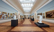 A large gallery space with a moulded ceiling with vaulted skylight and blue walls hung with many paintings. Sculptures and display cases stand on the wooden floor.