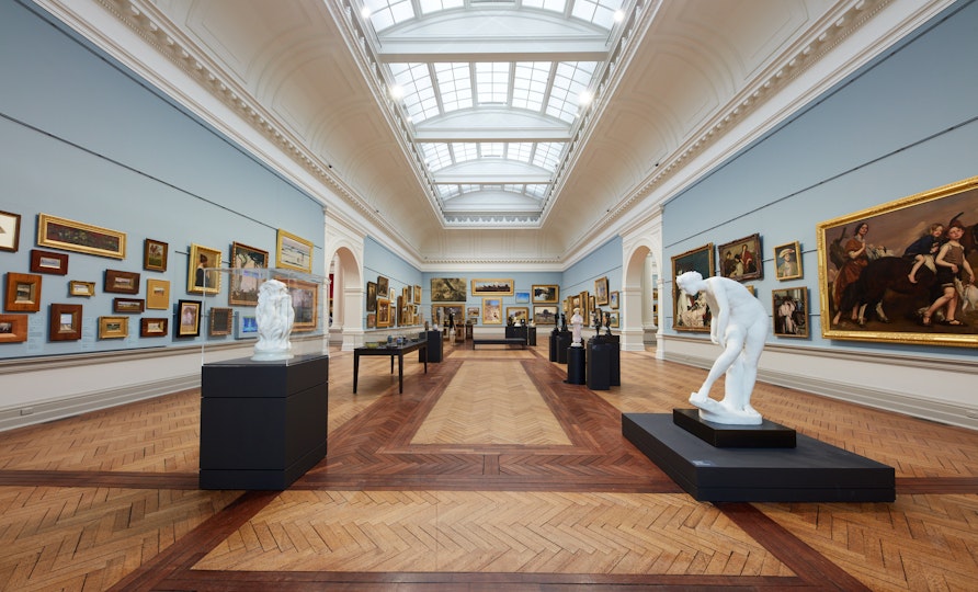 A large gallery space with a moulded ceiling with vaulted skylight and blue walls hung with many paintings. Sculptures and display cases stand on the wooden floor.