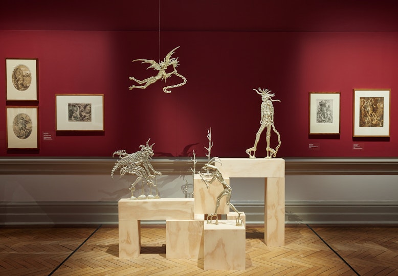 A red-walled gallery hung with framed etching in front of which are small sculptures of demon-like creatures