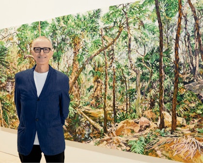 A person wearing glasses stands in front of large painting of bushland