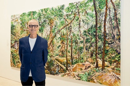 A person wearing glasses stands in front of large painting of bushland