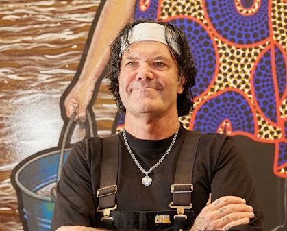 A person wearing a white headband, black long-sleeved t-shirt and black overalls stands with their arms crossed