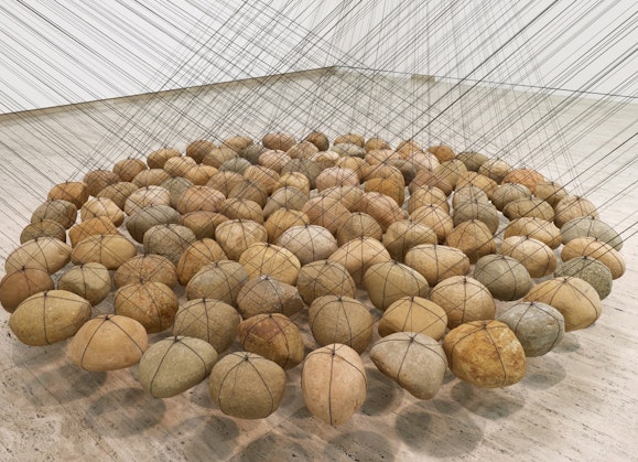Dozens of rounded stones of similar size suspended by strings in a tight grouping above the floor