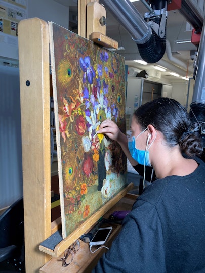 A person at an easel using applying a swab to the surface of a still-life painting of flowers