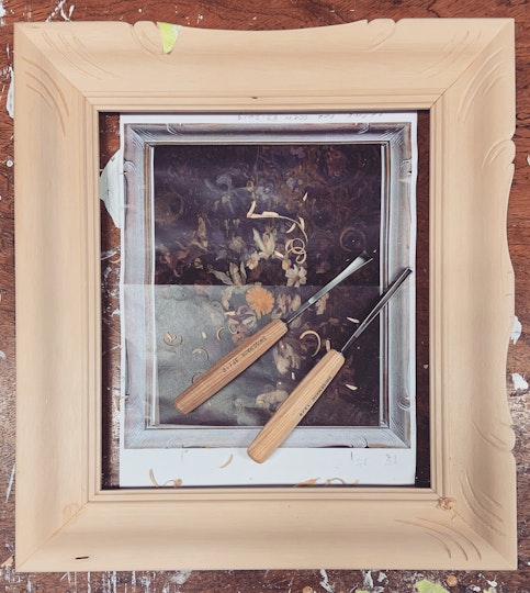 A partially carved, unpainted picture frame lies on a surface. Inside the frame are two chisels and a black-and-white image of a painting in a similar frame