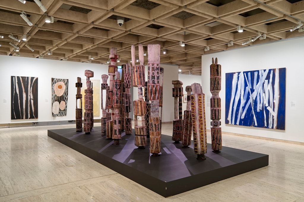 Installation view of the 20th-century galleries at the Art Gallery of New South Wales, featuring (foreground) Laurie Nelson Mungatopi, Bob Apuatimi, Jack Yarunga, Don Burakmadjua, Charlie Kwangdini and unknown artist Pukumani grave posts 1958; (walls, far left and far right) Tony Tuckson Five white lines (vertical), black ground 1970–73 and White lines (vertical) on ultramarine 1970–73; (wall, centre left) Noŋgirrŋa Marawili Baratjala – lightning and the rock 2018, photo © Art Gallery of New South Wales, Christopher Snee