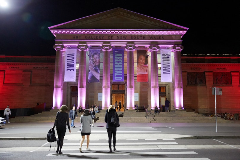 Visitors walking across the pedestrian crossing towards the entrance to the Gallery at night. The columns are uplit with pink lights.